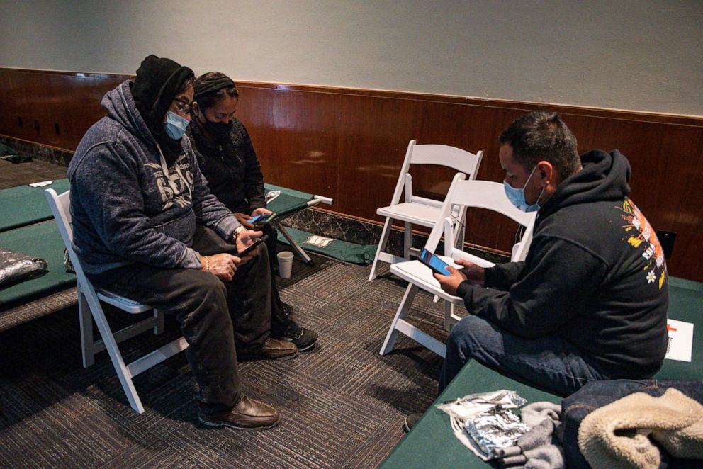 Miguel Mancilla and his wife, left, and Ramon Gutierrez, right, pass the time on their phones at a warming shelter inside the Copper Rose Building on Tuesday, Feb. 16, 2021, in Odessa, Texas. 